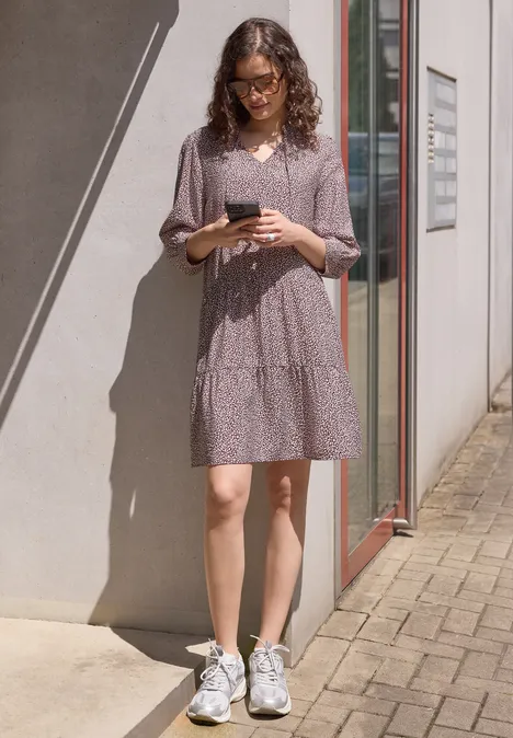 Knielanges Kleid mit V-Neck und Print vanilla sand Frau in lila gepunktetem Kleid und weißen Sneakern steht draußen an einer Hauswand und hält ein Handy.