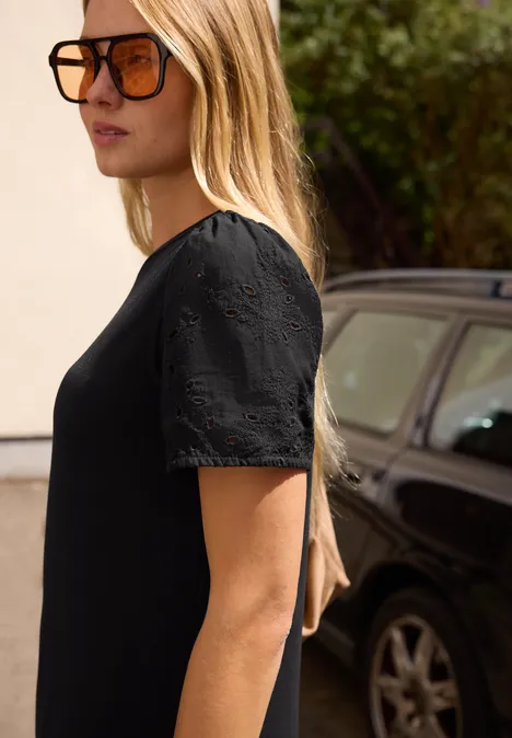 Eine Frau mit langen blonden Haaren in schwarzem Kleid steht seitlich vor einem geparkten schwarzen Auto.
