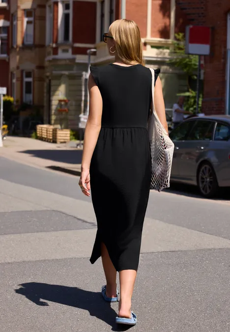 Femme en robe noire midi et baskets bleues marche dans une rue ensoleillée en tenant un sac en filet.
