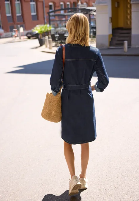Frau in jeansblauem Kleid geht mit Strohtasche über eine sonnige Straße.