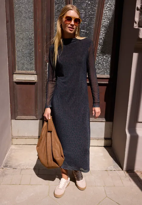 Junge Frau mit langem dunklem Kleid, weißen Sneakern und Sonnenbrille steht in sonniger Türöffnung und hält eine braune Tasche.