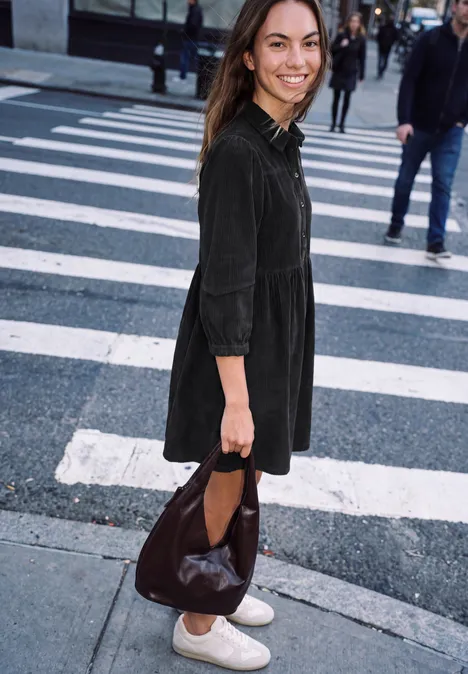 Kniehoge jurk van corduroy in effen kleur inked grey Glimlachende vrouw in zwarte jurk en witte sneakers steekt een stadsstraat over met een grote bruine tas.