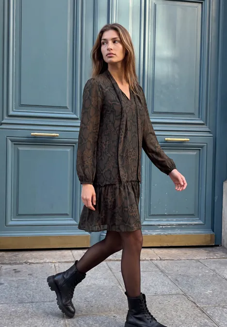 Knielanges Chiffonkleid mit Schalkragen charred brown Junge Frau in schwarzem, langärmligem Kleid und Boots steht vor einer blauen Tür auf dem Bürgersteig.