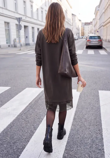 Minikleid mit Spitzendetail am Saum charred brown Frau in dunkelgrünem Kleid und Stiefeln, trägt Tasche und Kaffeebecher, geht über Zebrastreifen.