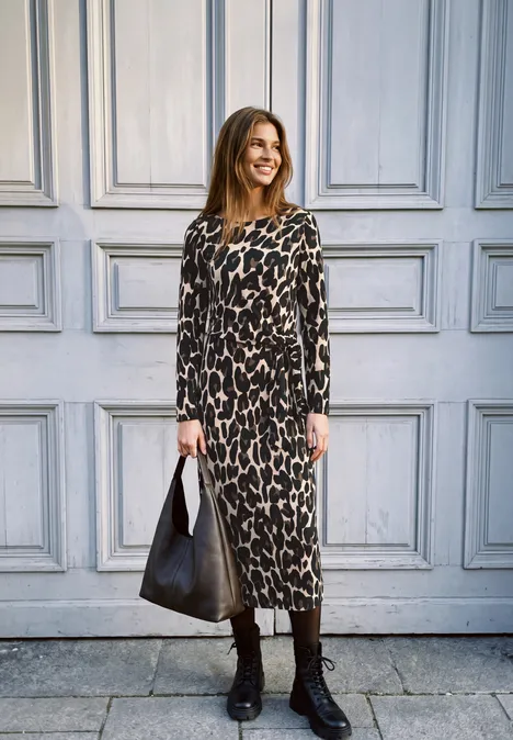 Robe mi-longue en jersey avec un nœud Black Femme en robe léopard longue devant une porte grise, avec bottes et sac noirs, souriante.