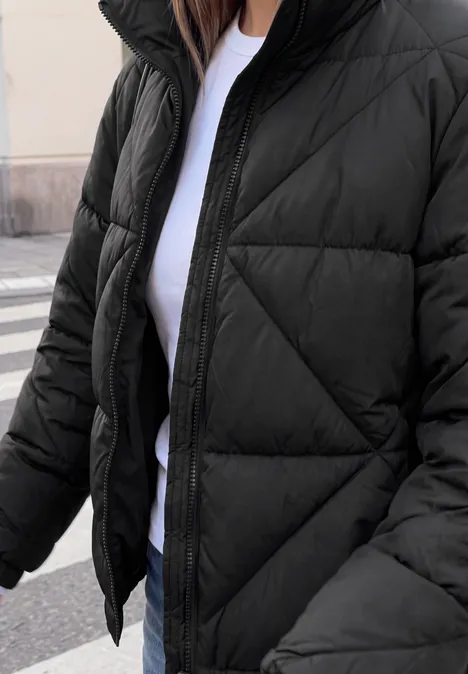 Person trägt schwarzen Steppmantel über weißem T-Shirt, dazu Jeans, steht auf Gehweg.