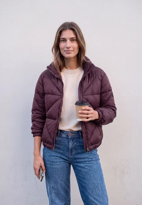 Jonge vrouw voor een witte muur, glimlacht, draagt paarse gewatteerde jas en jeans, met koffie en telefoon.