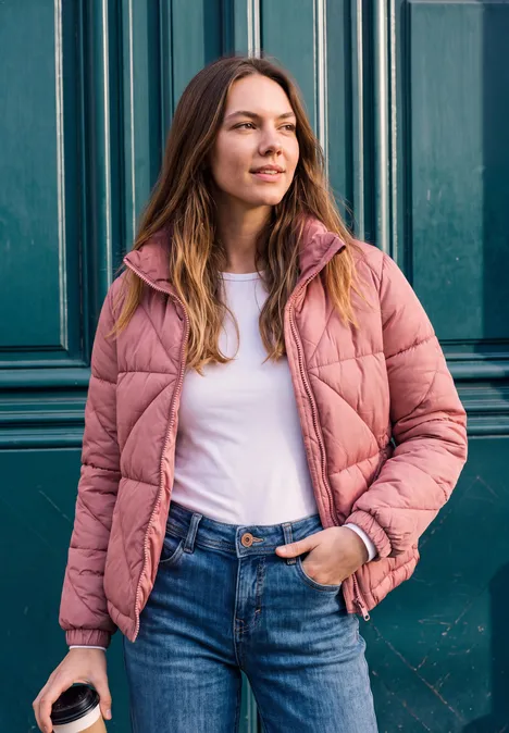 Kurze Jacke mit Stehkragen und Steppmuster Dark Vintage Rose Frau in rosa Steppjacke, weißem Shirt und Jeans hält Coffee-to-go vor grüner Metalltür.