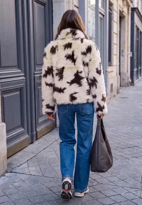 Vue de dos d'une femme en manteau blanc à étoiles noires, jean bleu et baskets, marchant en ville.