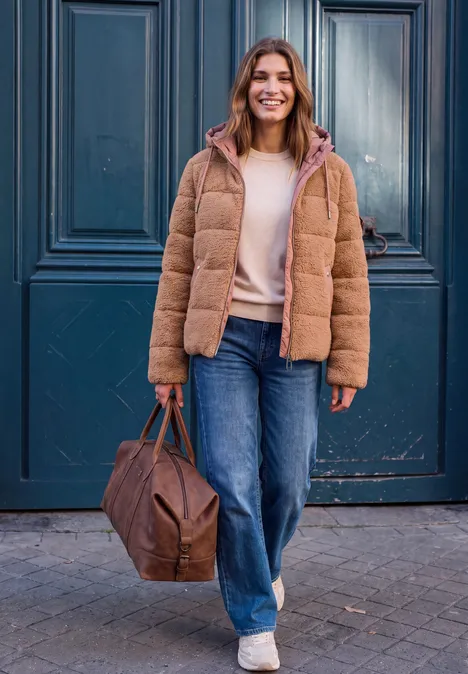 Veste en peluche à capuche avec des détails en softshell Honey Brown Eine Frau in brauner Steppjacke und Jeans trägt eine Tasche und lächelt vor einer blauen Tür.