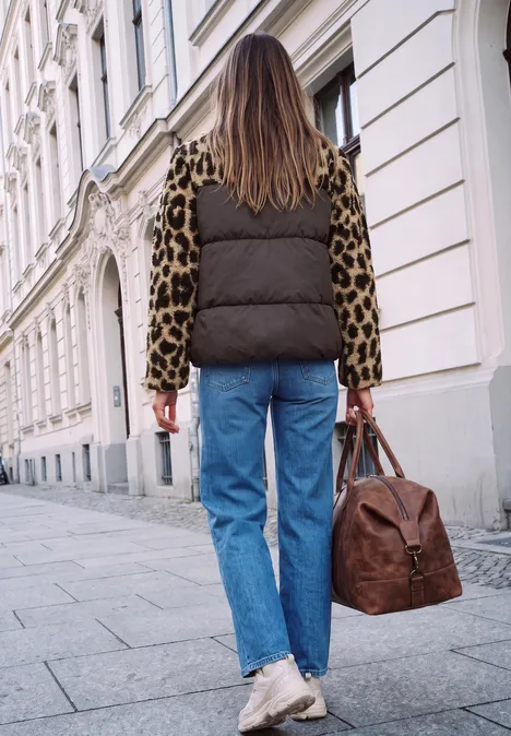 Une femme marche de dos en gilet matelassé, chemise léopard, jean et sac en cuir devant des immeubles.