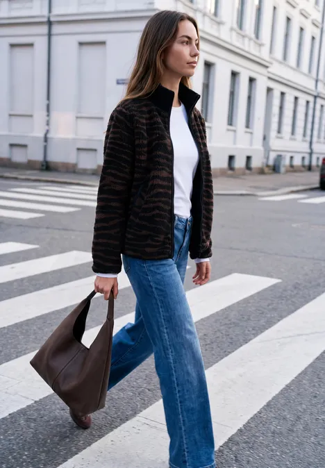 Une femme en jean, t-shirt blanc et veste zébrée traverse une rue urbaine en portant un sac marron.