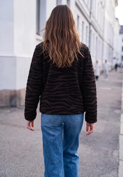 Une femme aux longs cheveux bruns, veste noire et jean bleu, marche dans une rue urbaine calme.