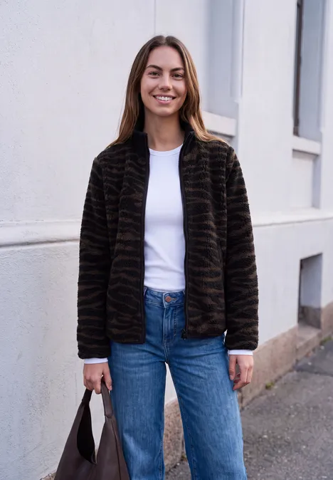 Une femme souriante en veste marron et jean est appuyée contre un mur, tenant un sac à main.