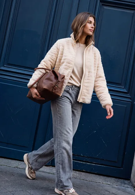 Une femme en jean, veste crème et sac à bandoulière marche devant une porte bleu foncé.