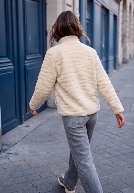 Femme en veste crème et pantalon gris marchant sur un trottoir pavé devant des portes bleues.