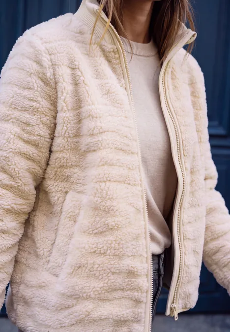 Veste teddy beige zippée, t-shirt clair en dessous, vue partielle du buste.