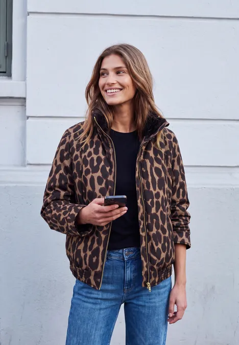 Junge Frau in Leopardenjacke hält ihr Smartphone und lächelt vor einer weißen Hauswand.