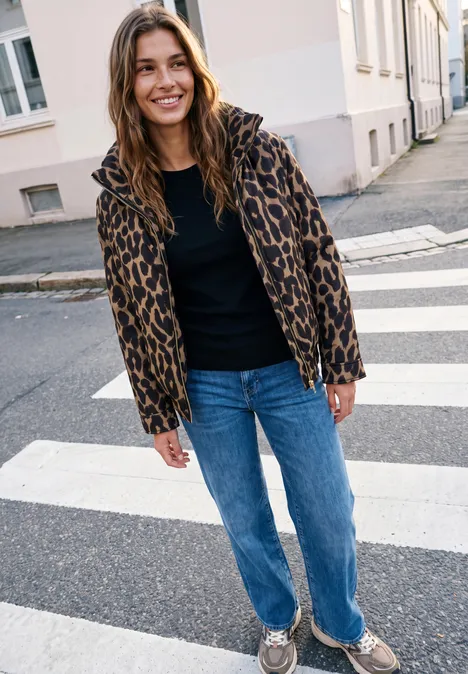 Eine lächelnde Frau mit leo-print Jacke, schwarzem Shirt, Jeans und Sneakern steht auf einem Zebrastreifen in der Stadt.