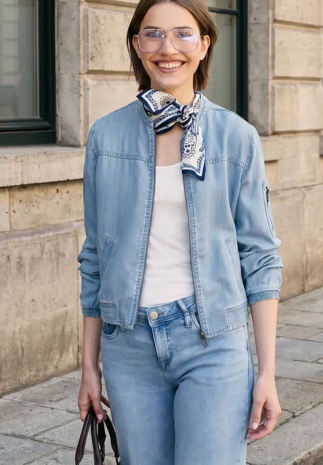 Vrouw in denimjack en jeans met witte top en sjaaltje, draagt handtas voor stedelijke gevel.