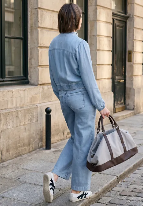 Persoon in lichtblauwe denim overall met grote tas loopt over een kasseienstraat langs een stenen gebouw.