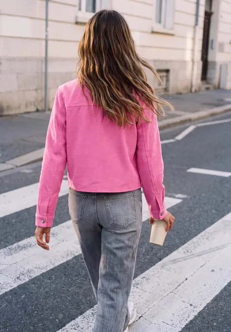 Frau mit pinker Jacke und grauer Hose überquert einen Zebrastreifen in einer Stadt, von hinten.