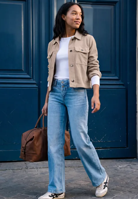 Eine junge Frau in Jeans, weißem Shirt und beiger Jacke trägt eine braune Tasche vor einer blauen Tür.
