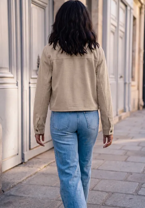 Frau von hinten in hellbeiger Jacke und blauen Jeans geht an einer Hauswand entlang.
