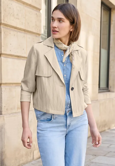 Vrouw in beige jas, spijkerbroek en sjaaltje voor een stenen muur.