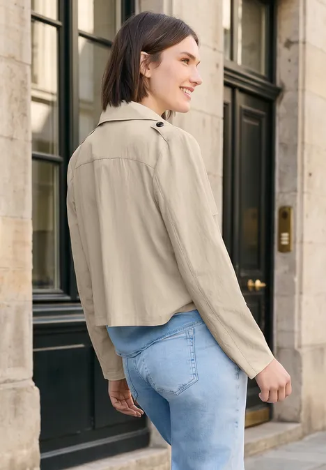 Een vrouw staat met haar rug naar de camera voor een ingang, ze draagt een korte beige jas en een blauwe jeans.