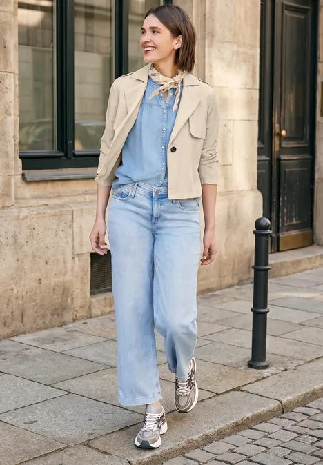 Vrouw in lichte spijkerbroek, blauw shirt, beige jasje en sneakers loopt op een geplaveid trottoir voor een gebouw.