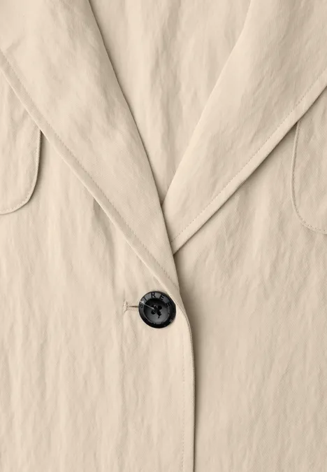Close-up van een beige blazer met revers en een zwarte knoop.