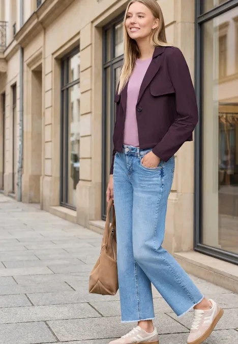 Frau in lila Blazer, rosa Top, weiter Jeans, Beuteltasche und Sneakern steht auf dem Bürgersteig vor Schaufenstern.