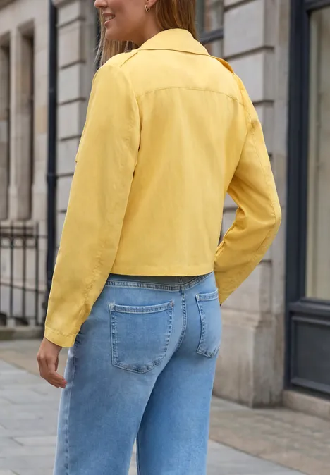 Frau von hinten in gelber Kurzjacke und blauen Jeans, stehend vor städtischer Kulisse.