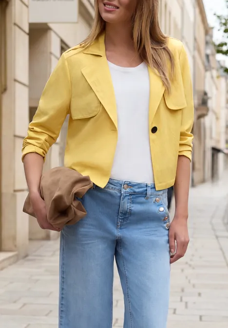 Frau in gelber Jacke, weißem Top und blauen Jeans, die einen Hut hält und auf einer gepflasterten Straße in der Stadt steht.