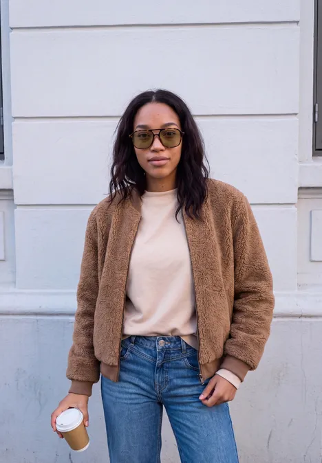 Frau mit brauner Teddyjacke, Jeans und Brille hält einen Kaffee und steht vor einer weißen Hausfassade.