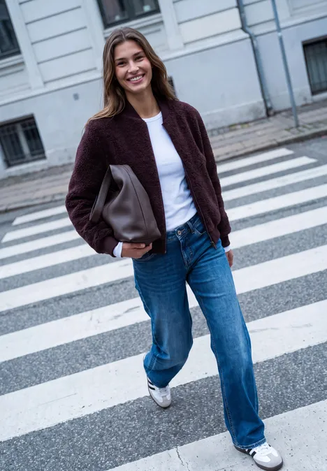 Teddyblouson mit Reißverschluss russet red Eine lächelnde Frau in Jeans, weißem T-Shirt und Jacke trägt eine Tasche, während sie über einen Zebrastreifen geht.