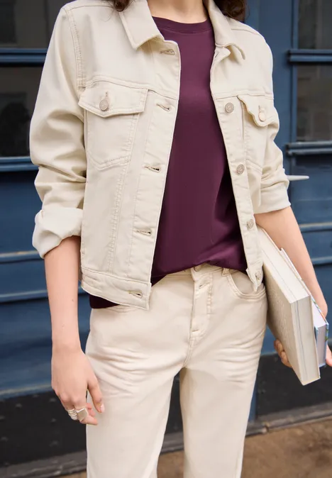 Eine Person trägt eine beige Jacke, lila Shirt und beige Hose, hält ein Buch.