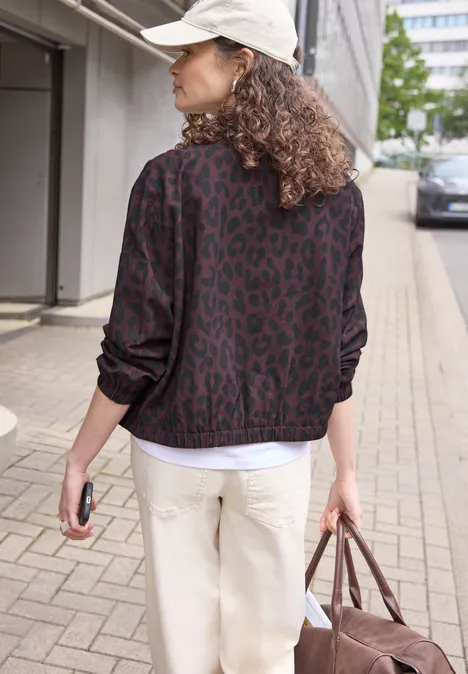 Rugaanzicht van een persoon met een patroonjas, witte pet en lichte broek, met een tas op het trottoir.