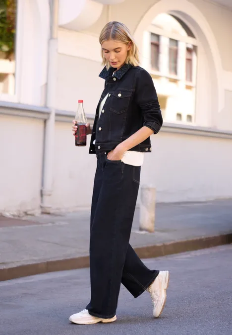 Frau in schwarzem Denim-Outfit und weißen Sneakern spaziert mit Getränk und Tasche an einer hellen Hausfassade.