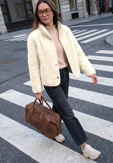 Frau trägt weiße Jacke, rosa Pullover, dunkle Jeans, weiße Sneaker, braune Reisetasche