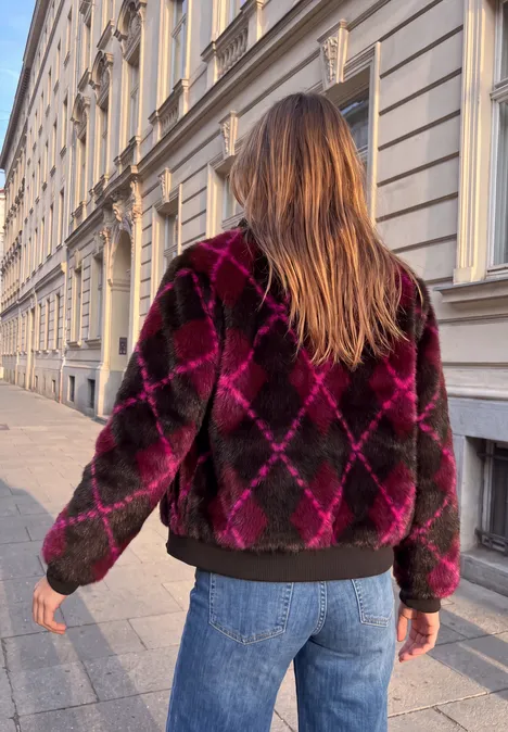 Eine Person mit langen Haaren trägt eine karierte, flauschige Jacke und Jeans und läuft eine Häuserzeile entlang.