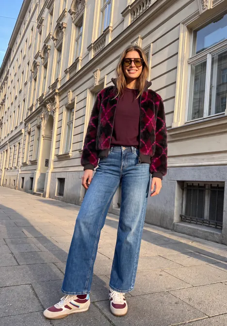 Frau in Jeans, burgunderroter Jacke und Sneakern steht entspannt vor historischer Fassade.