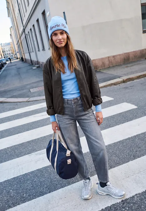 Blouson aspect cuir charred brown Junge Frau in Jeans, Bomberjacke und Mütze mit Tasche steht auf einem Zebrastreifen.