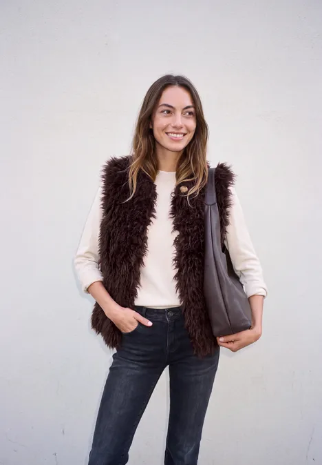 Lächelnde Frau in braunem Fellweste, hellem Pullover und Jeans, trägt eine graue Tasche vor hellem Hintergrund.