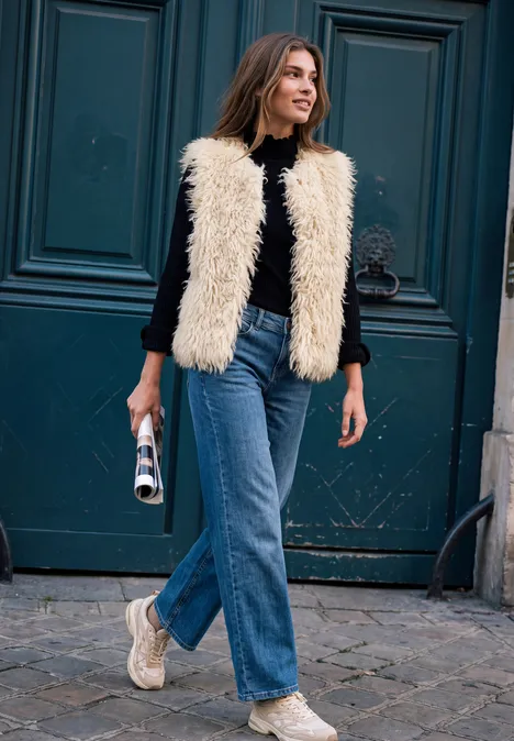 Eine junge Frau spaziert vor einer Tür, trägt schwarze Bluse, cremefarbene Fellweste, Jeans und weiße Sneaker.