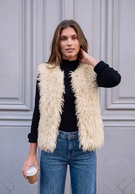 Frau mit hellbraunen Haaren, schwarzem Langarmshirt, cremefarbener Teddyweste und blauen Jeans hält Kaffee.