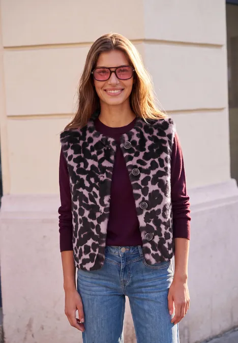 Lächelnde Frau mit Leopardmuster-Weste, dunkelrotem Oberteil, Jeans und Brille vor heller Hauswand.