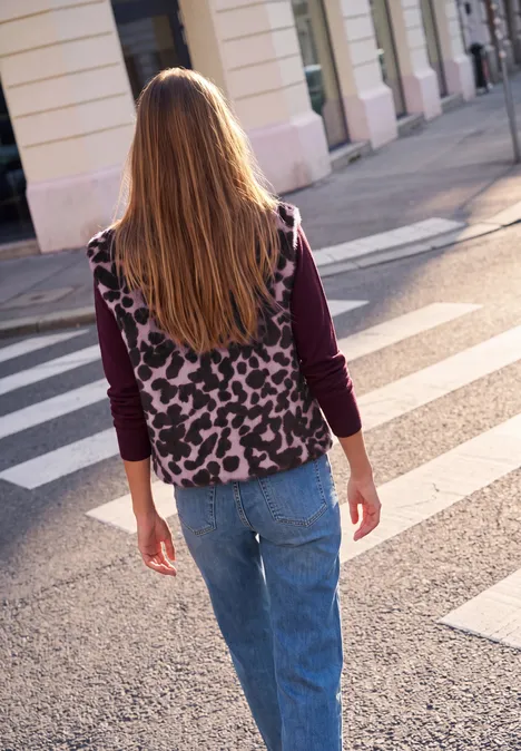 Frau mit langen blonden Haaren, gemusterter Weste und Jeans über einen Zebrastreifen in der Stadt.