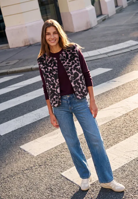 Eine junge Frau lächelt auf einem Zebrastreifen, trägt Jeans, weiße Sneakers und eine gemusterte Jacke.